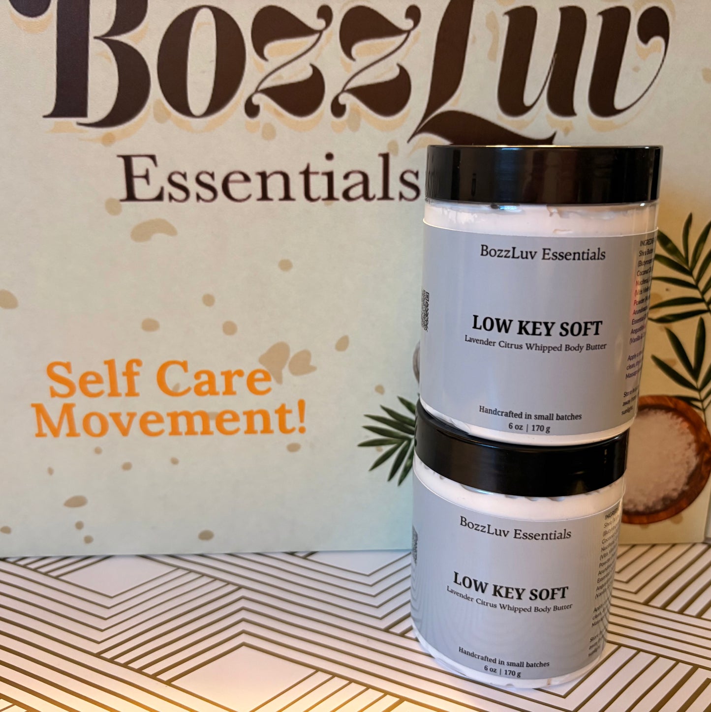Two jars of BozzLuv Essentials Low Key Soft lavender citrus whipped body butter stacked on a patterned surface with branded backdrop