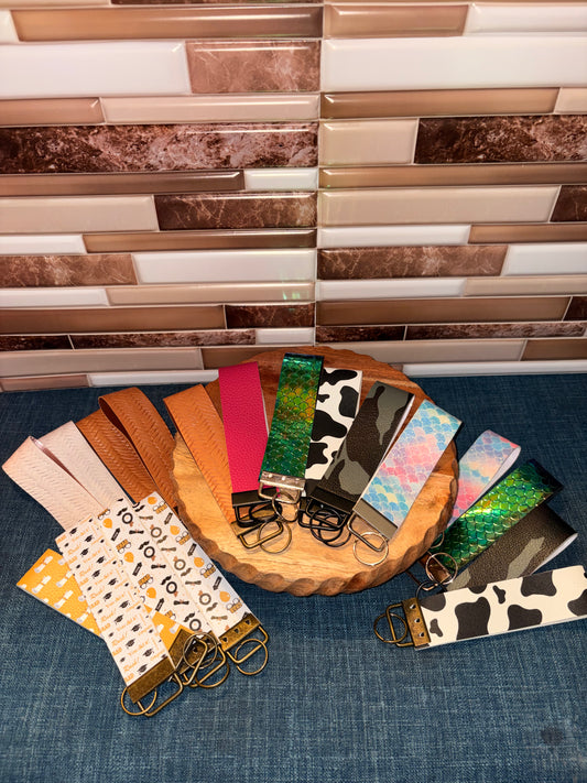 Group display of colorful faux leather wristlet keychains including cow print, shimmer, glitter, and solid color styles
