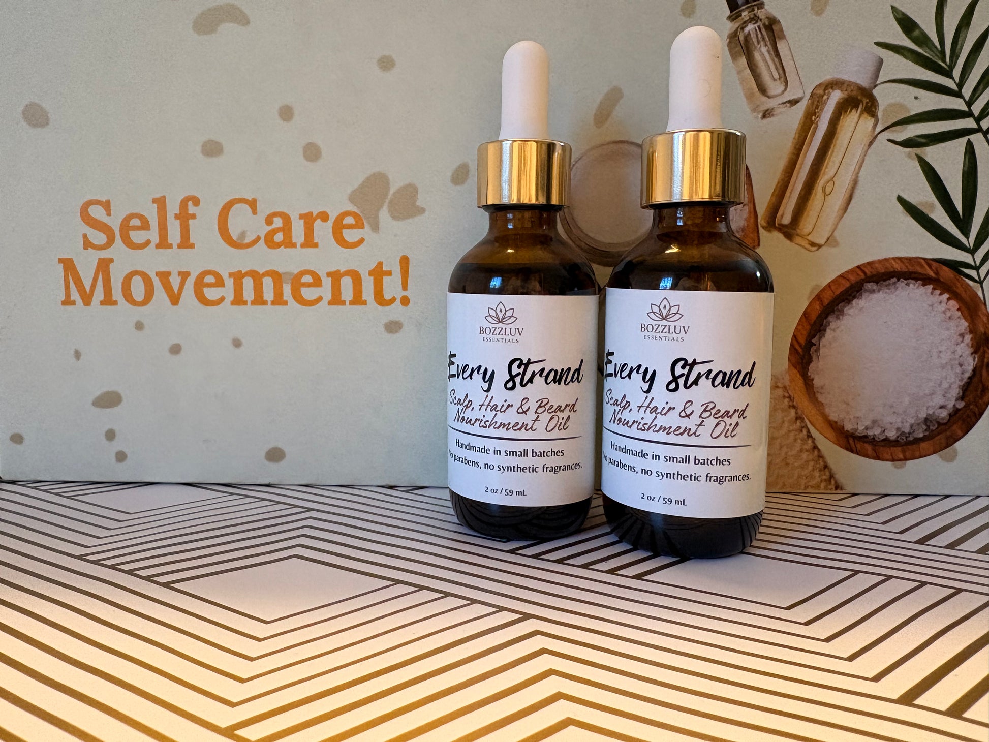Two 2 oz amber bottles of BozzLuv Essentials Every Strand Oil in front of branded “Self Care Movement” BozzLuv Essentials backdrop, positioned for lifestyle marketing photo.