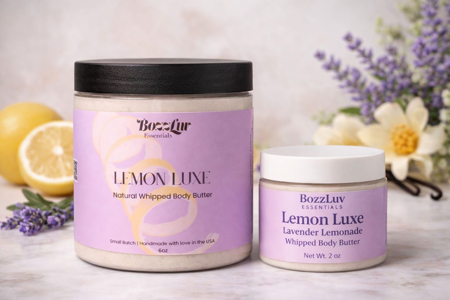 A styled product mockup showing a 6oz and a 2oz jar of BozzLuv Essentials Lemon Luxe Whipped Body Butter on a marble surface. Surrounded by fresh lemons, lavender sprigs, and vanilla flowers, the jars feature purple labels and black or white lids. The background is softly blurred for a fresh, spa-like feel.