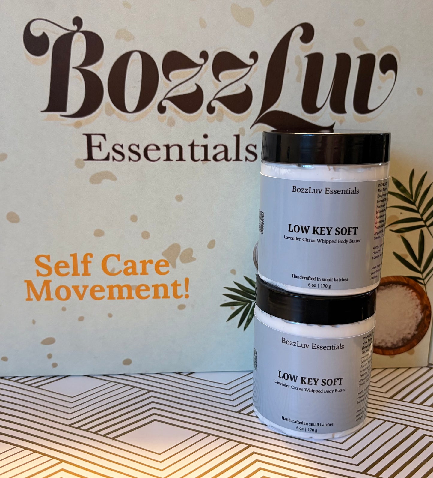 Two jars of BozzLuv Essentials Low Key Soft lavender citrus whipped body butter stacked on a patterned surface with branded backdrop