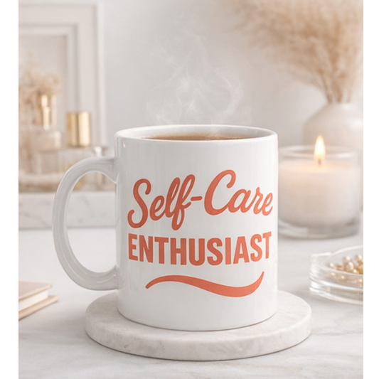 White mug with 'Self-Care Enthusiast' text on a marble coaster with candles and books in the background.