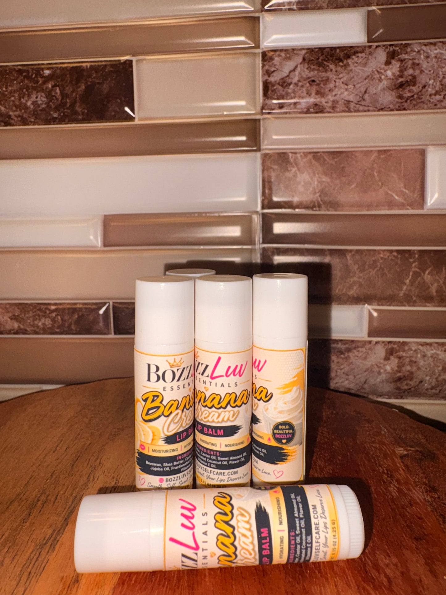 Three Banana Cream ice cream cans on a wooden surface with a tiled wall background. Set of Banana Cream lip balms by BozzLuv Essentials displayed together for lip care collection