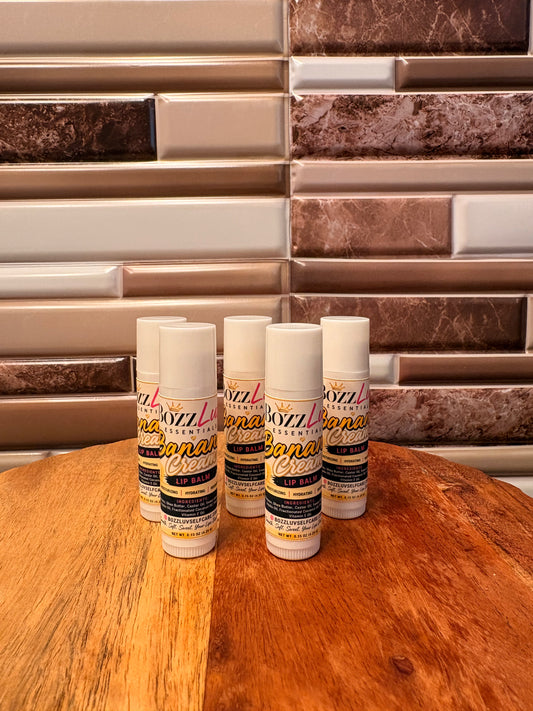 Five lip balms on a wooden surface with a tiled wall background. Close-up of BozzLuv Essentials Banana Cream Lip Balm label showing product details and ingredients