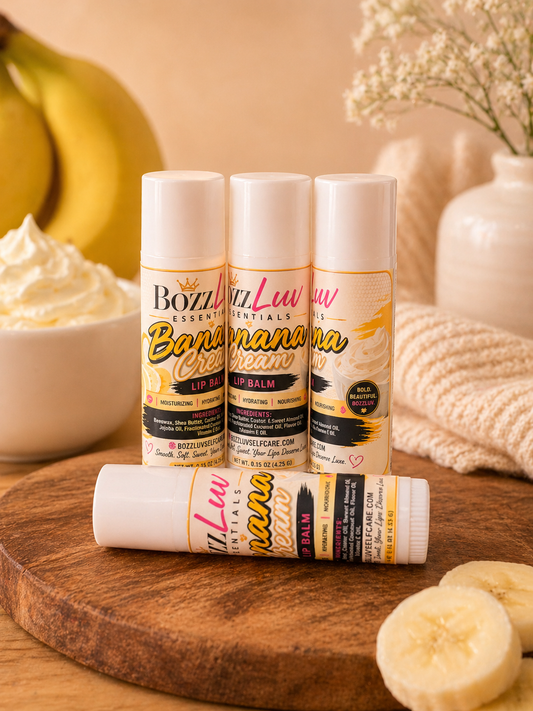 Three lip balm tubes labeled 'Banana Cream' on a wooden surface with bananas and whipped cream in the background. Banana Cream lip balm by BozzLuv Essentials styled with banana slices and cream for a soft, hydrating lip care experience