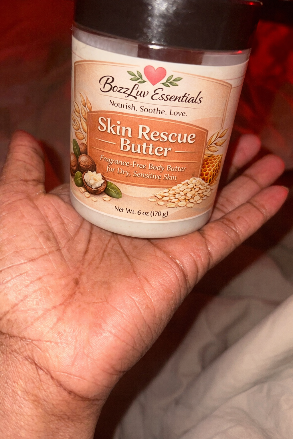 Hand holding a jar of Bozz Liv Essentials Skin Rescue Butter against a blurred background. BozzLuv Essentials Skin Rescue Butter 6 oz fragrance-free body butter for dry sensitive skin.