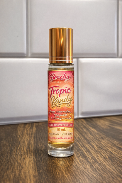 Roll-on perfume bottle labeled 'Tropic Kandy' by Bozz Luu on a wooden surface with a neutral background. BozzLuv Essentials Tropic Kandy roll-on perfume oil in a 10 mL glass bottle with gold cap and tropical label